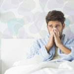 Sick man blowing his nose while sitting on bed at home