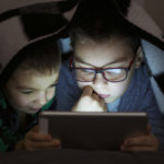 Two kids using tablet pc under blanket at night. Cute Brothers with tablet computer in a dark room smiling