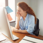 Portrait of tired sleepy businesswoman yawning, working at desk in the office in front of computer. Bored young female freelancer working on PC at home. Overwork and sleep deprivation concept