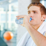 Refreshing after work out. Tired young man carrying towel on shoulders and drinking water while standing in gym
