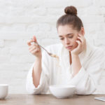 Young woman with no appetite, tired, bad mood and sleepy, sitting in the white loft kitchen, having unsavory unpalatable unappetizing breakfast