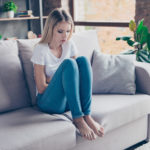 Upset woman has first symptoms of pms. She is sitting on a sofa and  touching her stomach. She is going to cry.