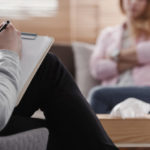 Back view of psychotherapist writing notes, assessing patient’s health and giving diagnosis to a woman sitting on a couch in the blurred background during counseling session