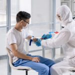 Young Chinese man in mask sitting in lab while doctor taking his blood