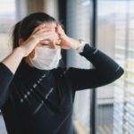Woman with headache and face masks indoors at home, Corona virus concept.