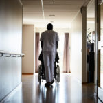 Full length rear view of male nurse pushing senior man on wheelchair at hospital corridor