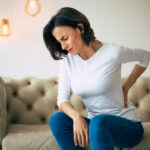 Axial pain. Close-up photo of a hurting woman, who is sitting on a couch and holding her lower back with her left hand.