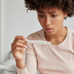 Embrarrassed woman with Afro hairstyle faces serious problem, dissatisfied with pregnancy test, dressed in pyjamas, has displeased expression, models indoor, thinks about abortion. How it possible?