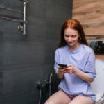 Cheerful redhaired young woman enjoying using phone sitting on toilet at bathroom, smiling looking to smartphone screen. Happy redhead female browsing internet