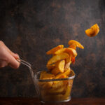 Side view fried potatoes with human hand in grid for deep frying on stone background