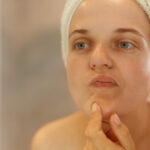 Closeup portrait of beautiful woman looking at her face, trying find acne, posing with naked shoulders and towel over head, doing morning beauty procedures.