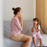 Physiotherapist wearing striped casual shirt working on speech defects or difficulties with small child girl at home while sitting on sofa, private lessons for improving sounds pronunciations.