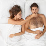 Irritated woman covers nose of husband, cannot fall asleep because of loud snoring, disturbed by noise. Spouses lie in snow white bed together. Unpleasant awakening, insomnia, sleep disorder.