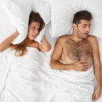 Irritated woman blocks ears, covers by pillow, looks with anger at snoring husband, cannot fall asleep, feels annoyed, has problems with sleeping, lie in white bed. Man has problems of sleep apnea