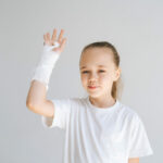 Medium shot portrait of cheerful little girl with broken hand wrapped in white plaster bandage greeting lifting arm looking at camera.