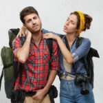 Attractive young European female with photo camera massaging neck of her frustrated sick boyfriend who is suffering from concussion after long hard hiking trip in mountains. People and traveling