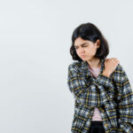 preteen girl holding hand on shoulder in shirt,checked jacket and looking painful , front view. space for text