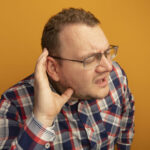 man in glasses and checked shirt with hand over ear listening to gossips standing over orange background