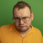 man in glasses and orange shirt looking at camera dissatisfied frowning eyebrows standing over green background