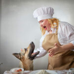 A fat funny female cook in a hat and apron in the kitchen with big shepherd dog. Dog steals food, cook swears. Body positive