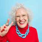 Portrait of cheerful nice looking elderly woman smiles happily makes peace gesture shows v sign dressed in red jumper with necklace expresses positive emotions isolated over blue background.