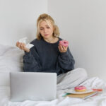 Portrait of sad, crying young woman, staying at home, sitting in bed with doughnut and comfort food, looking at something upsetting on laptop screen